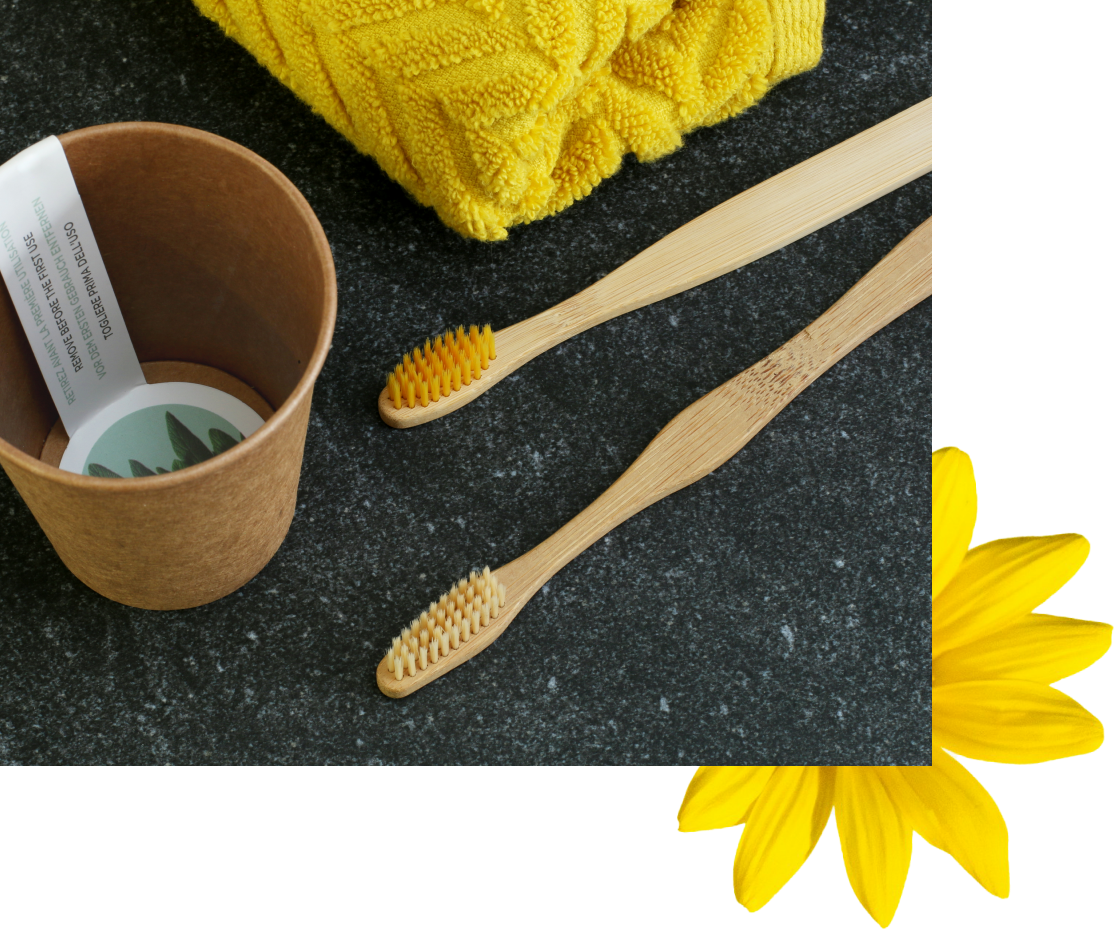Ein Paar Bambuszahnbürsten liegt neben einem braunen Papierbehälter, mit einem leuchtend gelben Handtuch und einer Margerite in der Ecke. Die Szene hat eine klare, minimalistische Ästhetik.