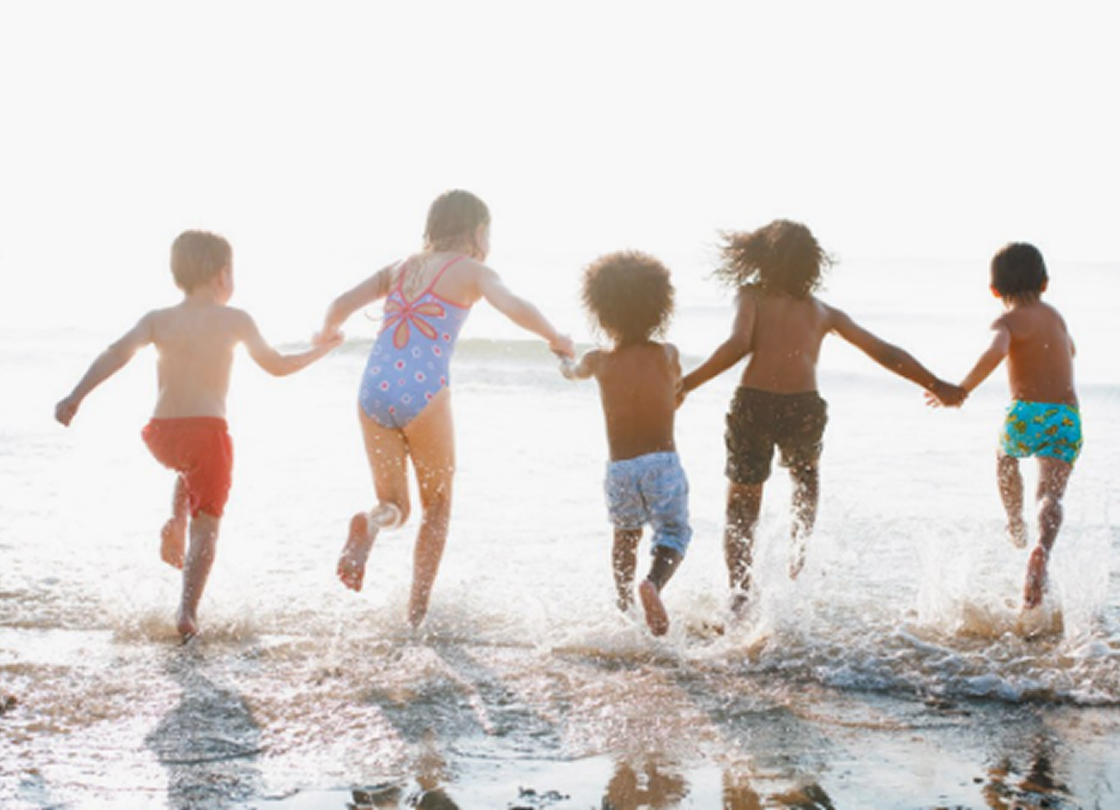 Eine Gruppe von fünf Kindern, die sich an den Händen halten, watet fröhlich ins Meer hinein und spritzt Wasser, während sie am Ufer entlang rennt. Die Sonne scheint ihnen in den Rücken und schafft eine helle, fröhliche Atmosphäre.