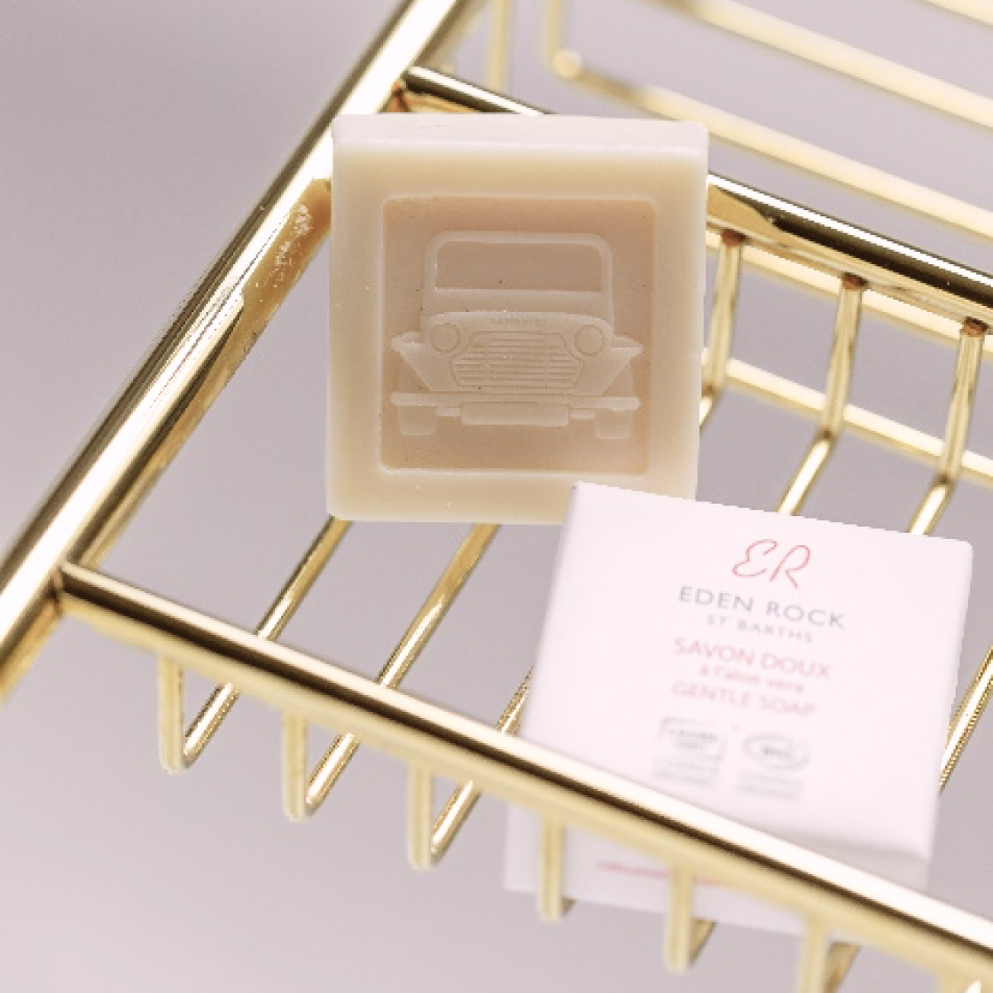 Ein Stück helle Seife mit einem Oldtimer-Motiv liegt auf einer goldenen Seifenschale neben einer kleinen weißen Schachtel mit der Aufschrift „Eden Rock“ – eine milde Seife.
