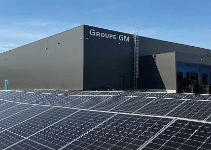 Um edifício moderno com a placa «Groupe GM» na fachada, com um telhado coberto de painéis solares sob um céu azul claro.