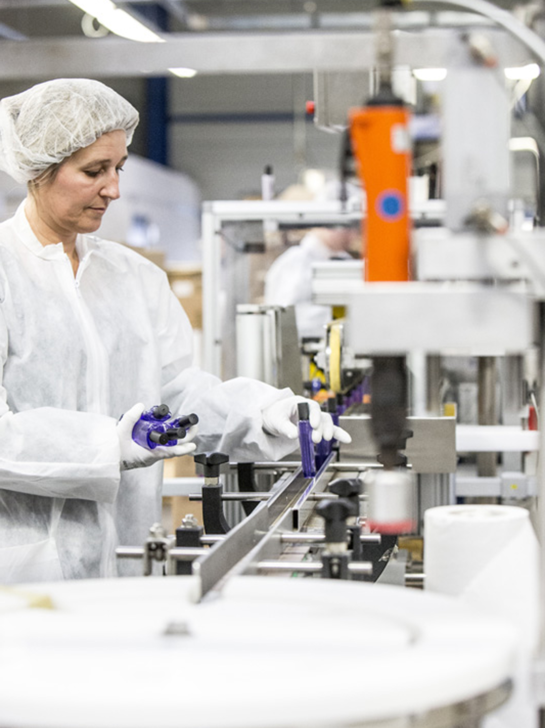 Eine Arbeiterin in einem weißen Laborkittel und einer Haube bedient Maschinen in einer Produktionsstätte und bearbeitet ein violettes Bauteil am Fließband.