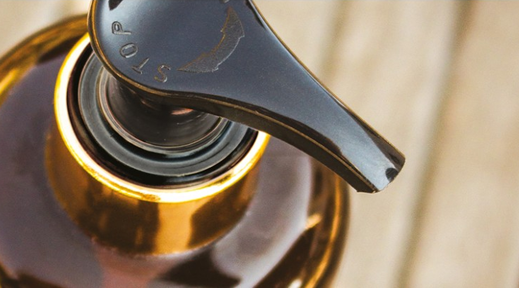Close-up de um dispensador preto com bomba em uma garrafa de vidro, com uma abertura circular e uma superfície de madeira ao fundo.
