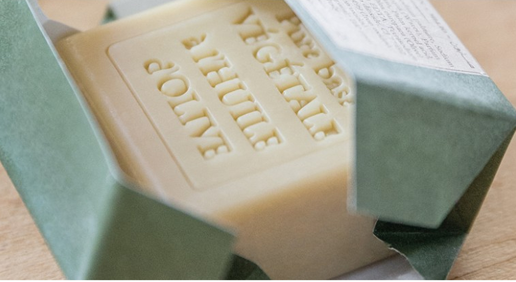 Uma barra de sabonete vegetal parcialmente desembrulhada de uma embalagem verde, com texto em relevo visível na superfície do sabonete.