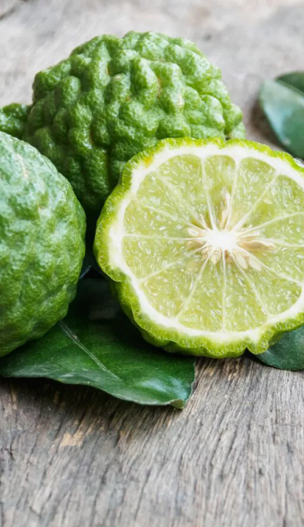 Nahaufnahme einer ganzen und halbierten Bergamotte-Limette auf einer Holzoberfläche, mit grünen Blättern im Hintergrund. Die Frucht hat eine unebene, grüne Schale und ein hellgelb-grünes Fruchtfleisch mit Kernen.
