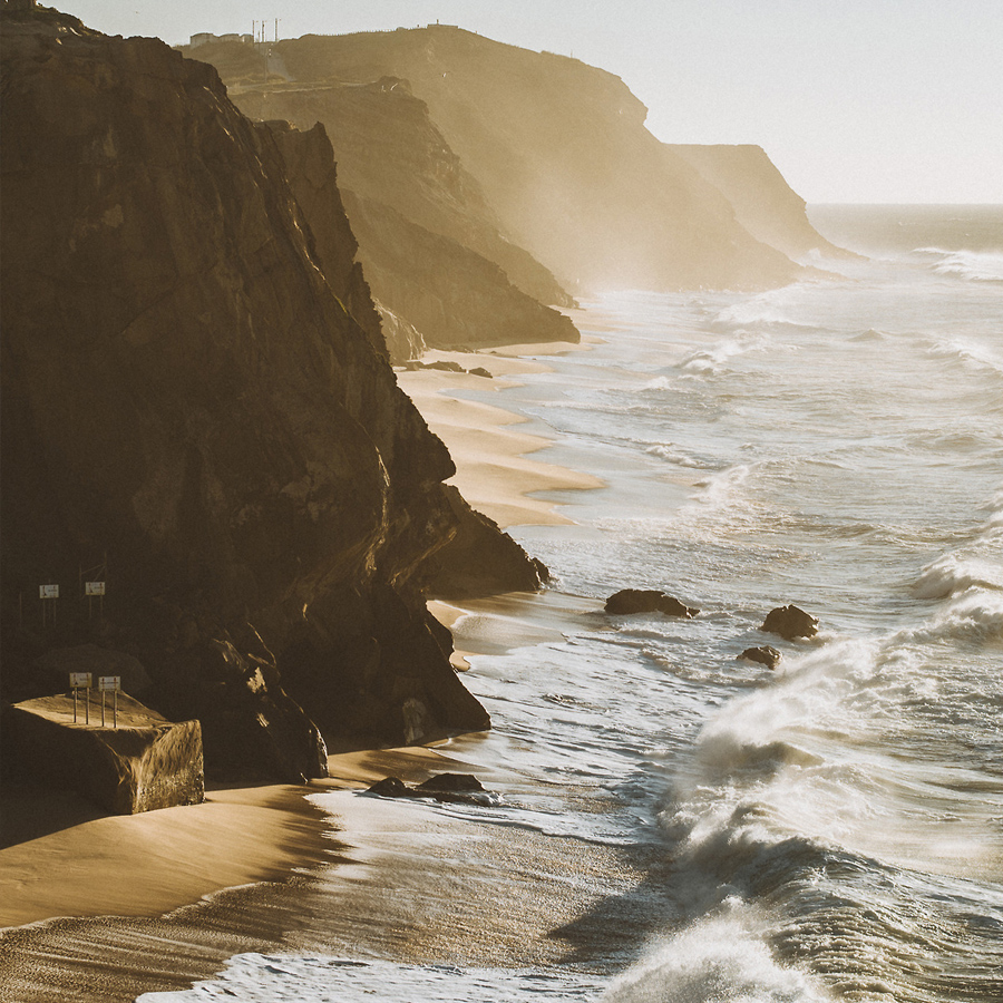 Una costa frastagliata con ripide scogliere si affaccia sull'oceano, dove le onde si infrangono sulla riva in un'atmosfera avvolta dalla nebbia e illuminata dal sole. La scena cattura la bellezza naturale e la potenza dei paesaggi costieri.