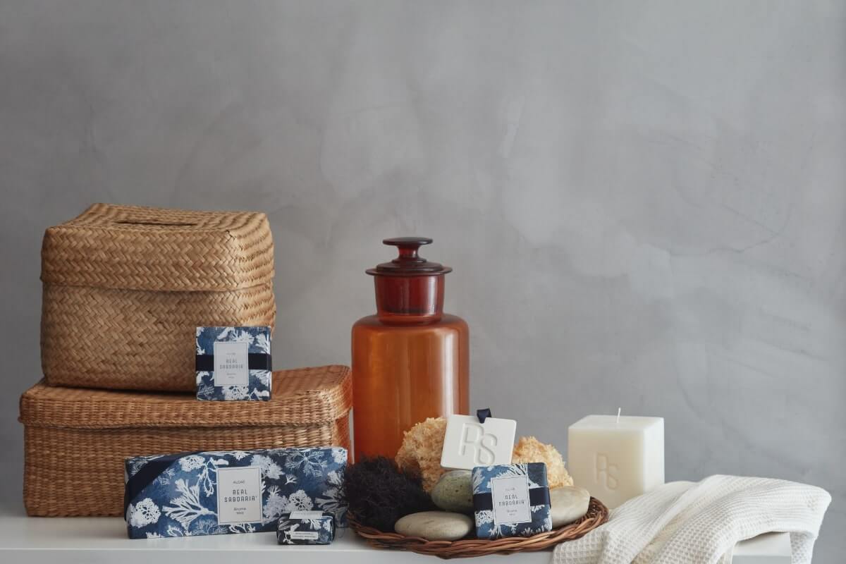 Una collezione di prodotti da bagno naturali e oggetti decorativi disposti su uno sfondo neutro, tra cui cesti intrecciati, un barattolo di vetro, una candela e saponette con confezioni floreali.
