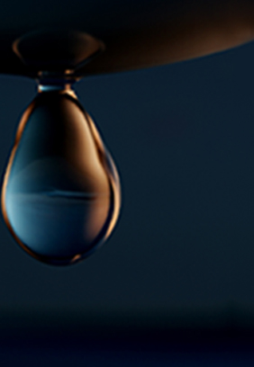 Ein einzelner Wassertropfen hängt an einer Oberfläche, reflektiert einen verschwommenen blauen Hintergrund und wird von sanftem Licht beleuchtet.