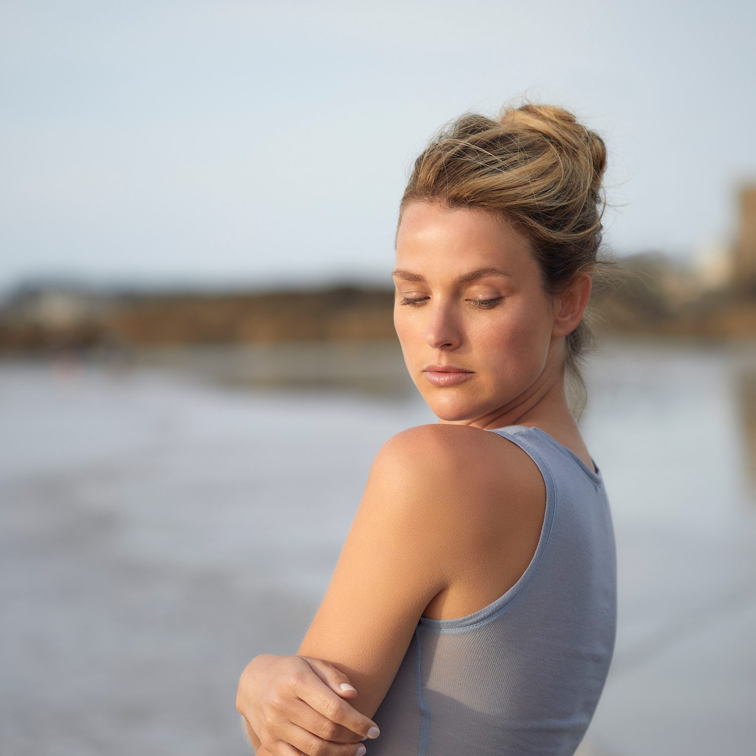 Een vrouw met een knot staat aan het strand; ze kijkt vredig met haar ogen dicht en draagt een grijze tanktop. De zee en het zachte zand zijn wazig op de achtergrond.