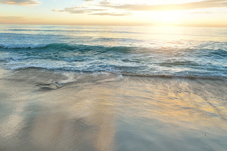 Eine ruhige Strandszene bei Sonnenuntergang, mit sanften Wellen, die an die Küste schlagen, und einem sanften Schimmer, der sich auf dem nassen Sand widerspiegelt.