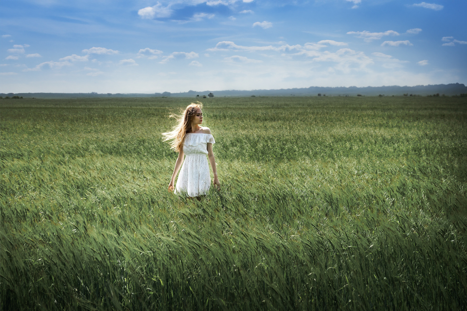 Eine junge Frau in einem weißen, schulterfreien Kleid steht inmitten von hohem grünem Gras, umgeben von einem weiten offenen Feld unter einem blauen Himmel mit vereinzelten Wolken. Ihr Haar weht sanft im Wind.