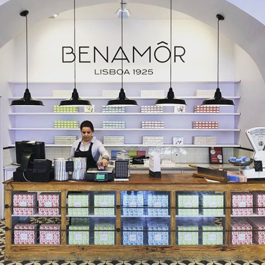 Un negozio dall'arredamento curato, con un bancone in legno e scaffali che espongono scatole colorate di prodotti, con una donna alla cassa e il marchio "BENAMÔR" ben visibile sulla parete.