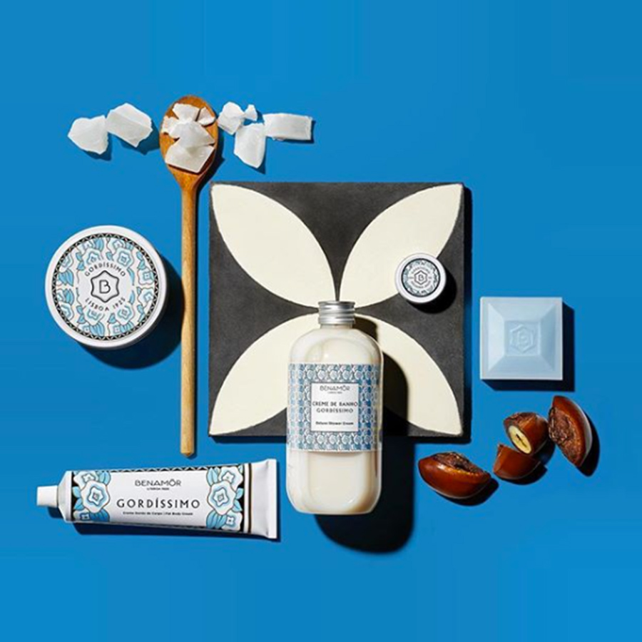 Uma variedade de produtos de beleza dispostos sobre um fundo azul brilhante, incluindo um frasco de loção, um tubo de creme, um sabonete e elementos decorativos, como uma colher de madeira e pedaços de coco.