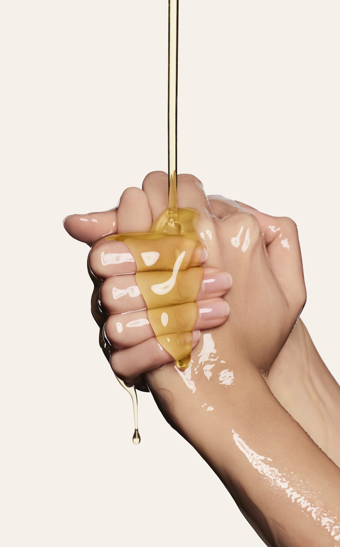 Nahaufnahme von zwei Händen, die ein tropfendes Stück Honig oder eine goldene, zähflüssige Flüssigkeit halten, wobei die Textur und der Glanz betont werden. Der Hintergrund ist in einer sanften, neutralen Farbe gehalten.