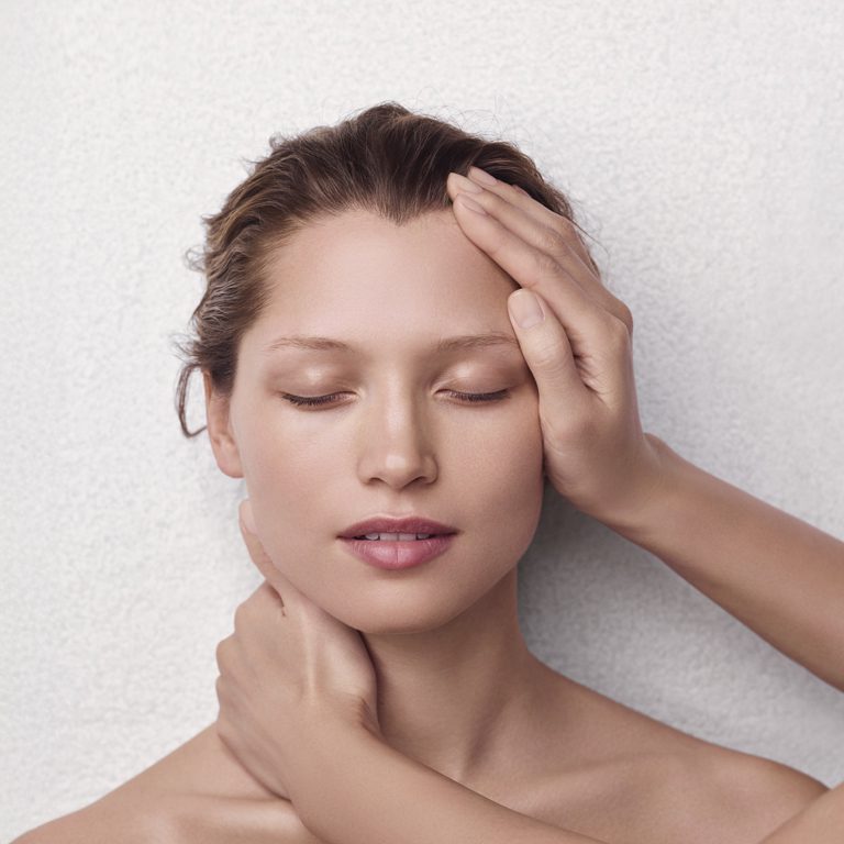 Eine ruhige Frau mit geschlossenen Augen und dezentem Make-up lässt sich vor einem sanften, neutralen Hintergrund sanft das Gesicht streicheln.
