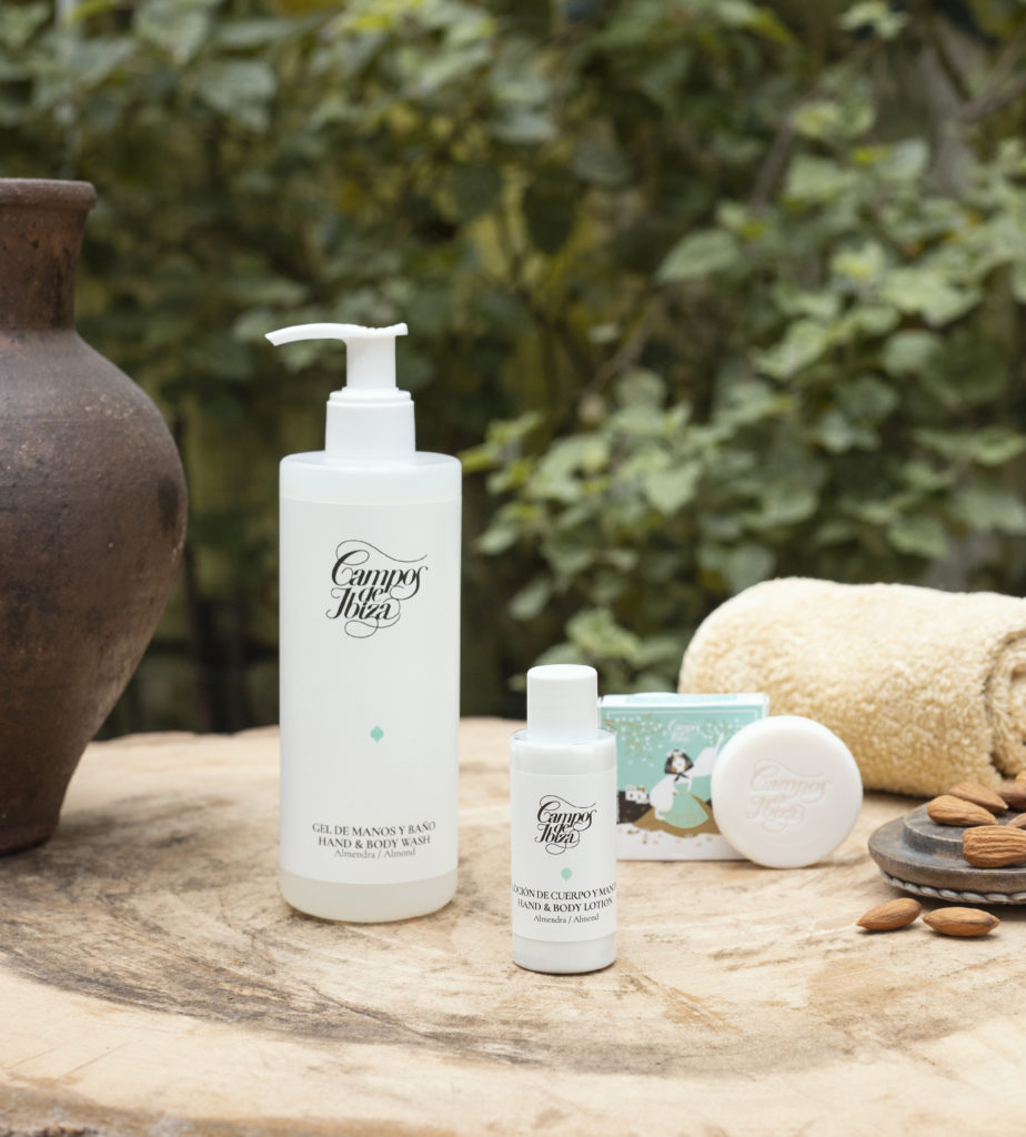 Una collezione di prodotti di bellezza, tra cui un flacone con dosatore grande, un flacone piccolo e una saponetta, esposti su un tavolo di legno con un vaso di terracotta e mandorle sullo sfondo. Il fogliame verde crea un ambiente naturale.