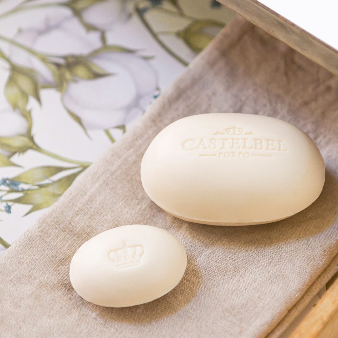 Dois sabonetes de cor creme estão dispostos sobre um tecido neutro, sendo que um deles, maior, exibe a inscrição em relevo «CASTELBEL PORTO» e o outro, menor, apresenta um desenho de coroa. O fundo inclui um padrão floral.