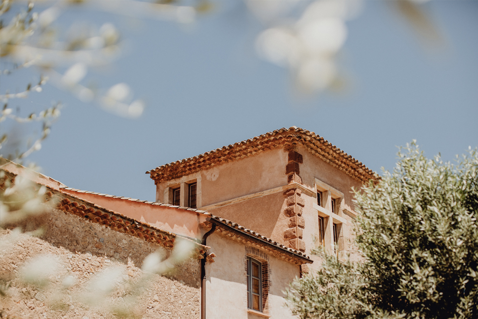 Una vista ravvicinata di un edificio rustico con tetto in terracotta, parzialmente incorniciato da rami di ulivo contro un cielo azzurro e limpido.