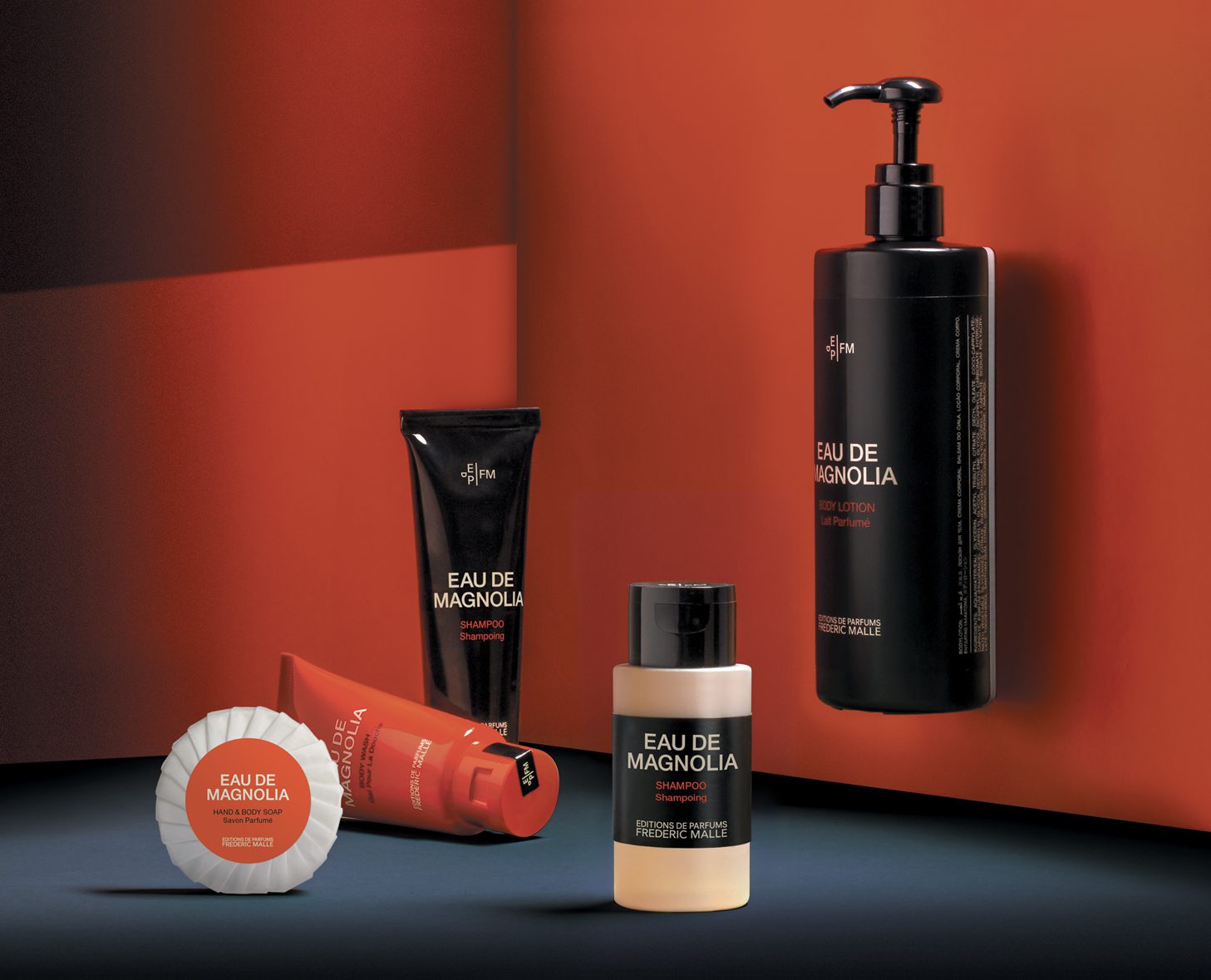 Una collezione di prodotti per la cura personale Eau de Magnolia esposta su uno sfondo rosso e nero a contrasto, che include una lozione per il corpo, uno shampoo e una crema.
