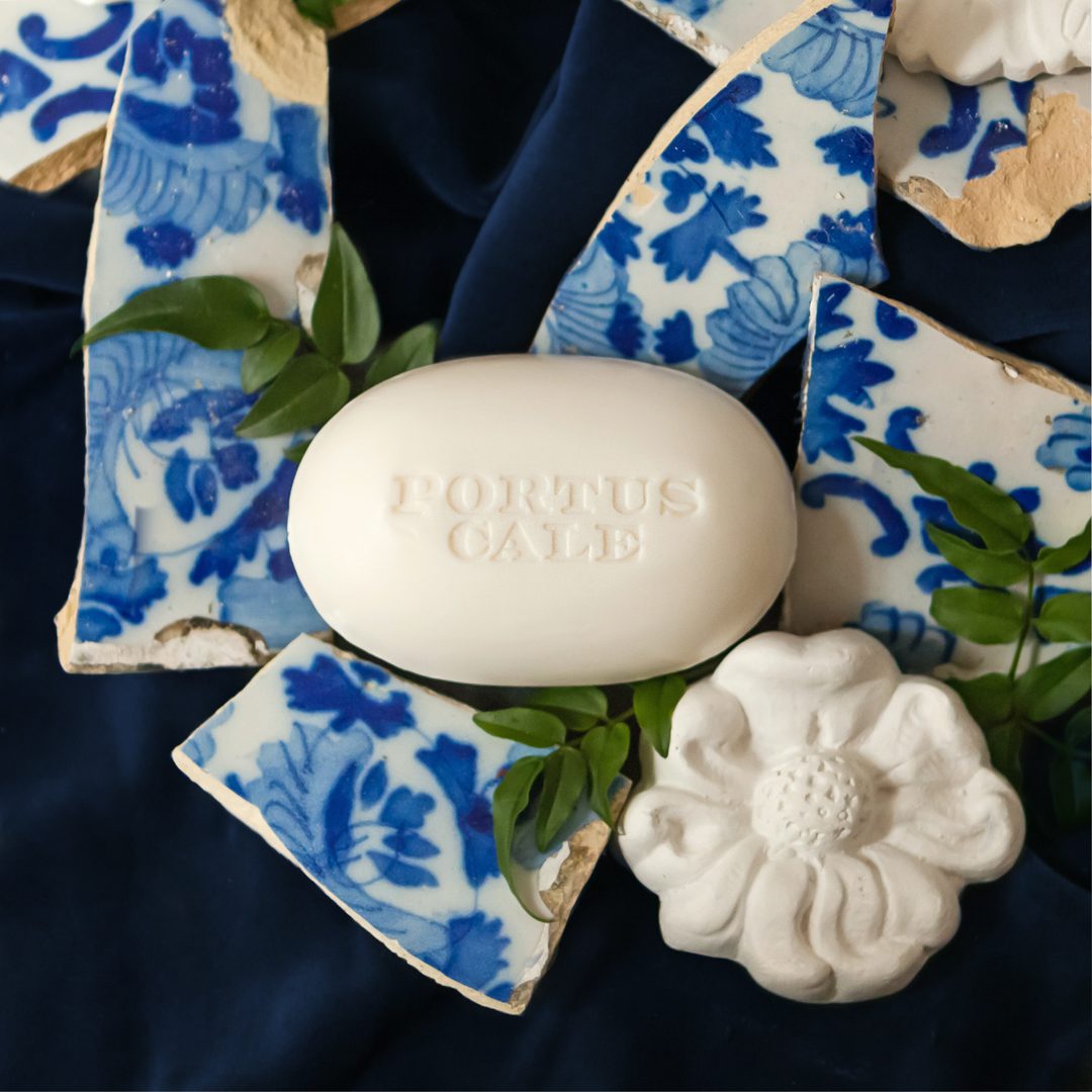 Una saponetta bianca con la scritta "PORTUS CALE" in rilievo, circondata da frammenti di ceramica blu e bianca e da una scultura floreale bianca su uno sfondo di tessuto scuro.