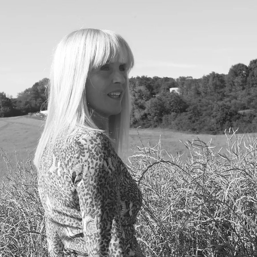 Uma mulher de cabelo loiro comprido está num campo, de perfil, com vegetação e árvores ao fundo. Ela veste um vestido estampado e parece estar a sorrir.