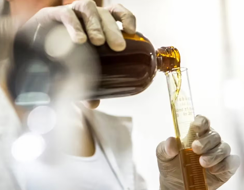 Um técnico de laboratório com luvas despeja um líquido castanho de uma garrafa para um cilindro de vidro graduado. O fundo está desfocado, com o foco na ação de despejar.