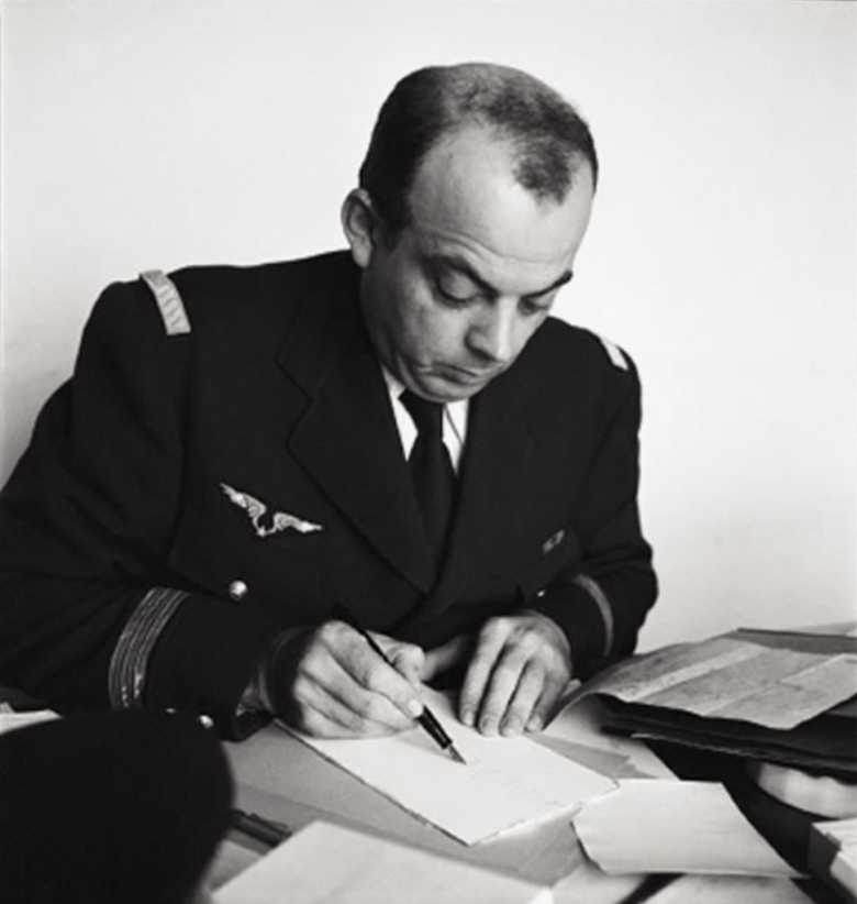 Ein Mann in Militäruniform sitzt an einem Schreibtisch und schreibt konzentriert auf ein Blatt Papier, um ihn herum liegen verstreut Dokumente.