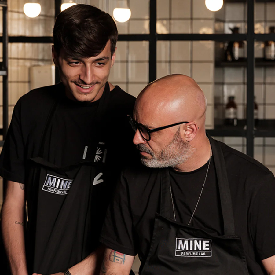 Dois homens com aventais pretos estão num ambiente de laboratório, a sorrir e a conversar. No fundo, vêem-se prateleiras com recipientes de vidro e uma iluminação acolhedora.