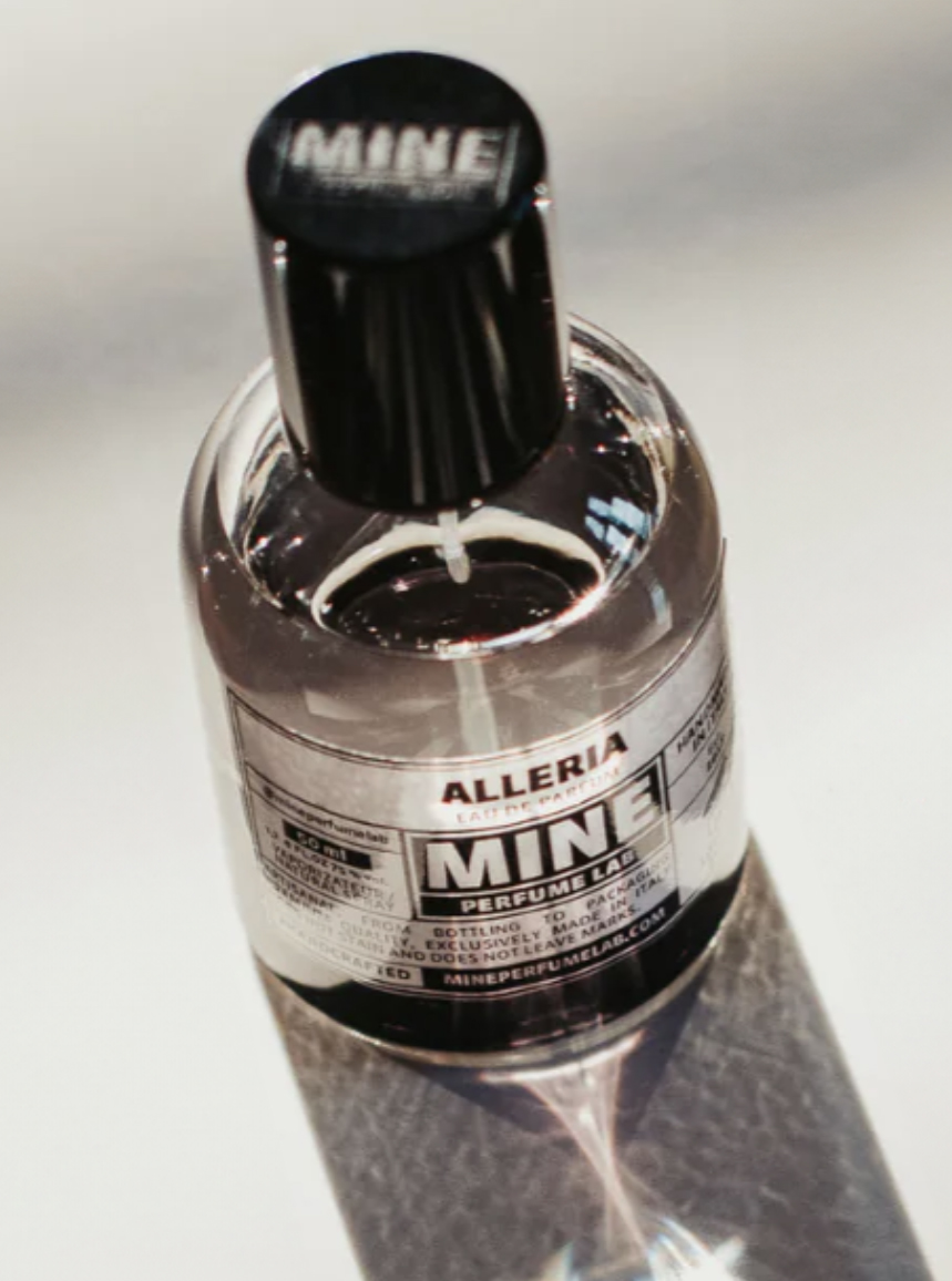 frascos de perfume transparente frascos uma tampa preta e um rótulo com as palavras «MINE» e «ALLERIA». O frascos posicionado de forma a criar uma sombra sobre uma superfície refletora.
