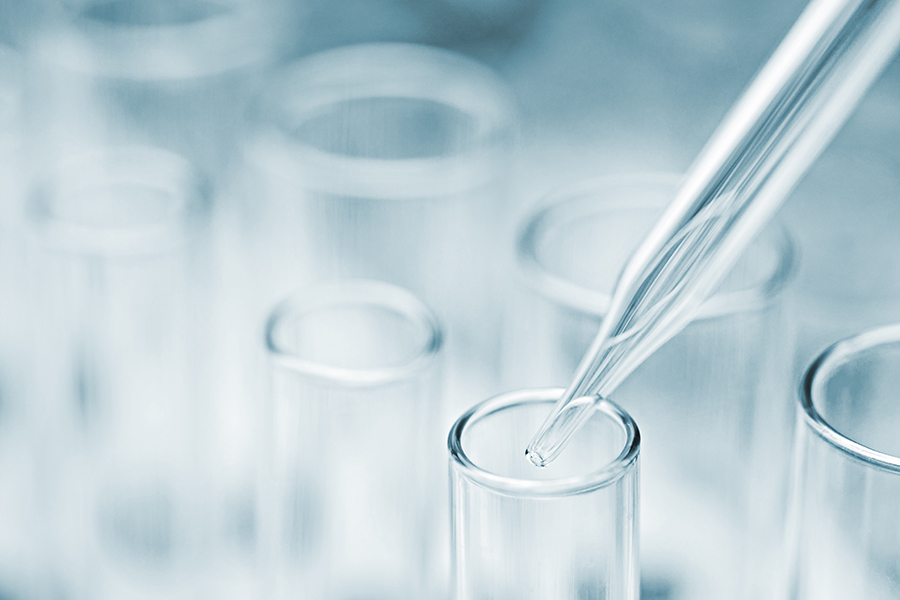 Nahaufnahme einer Pipette, die über einem durchsichtigen Reagenzglas schwebt, vor einem unscharfen Hintergrund mit weiteren Reagenzgläsern. Das Bild hat einen kühlen Blaustich.