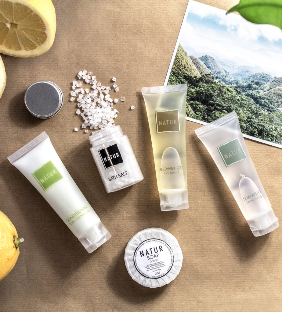 Una composizione di prodotti per la cura personale, tra cui tubetti di balsamo, bagnoschiuma e shampoo, un barattolo di sali da bagno e una saponetta, il tutto su uno sfondo di montagne lussureggianti e accompagnato da limoni freschi.
