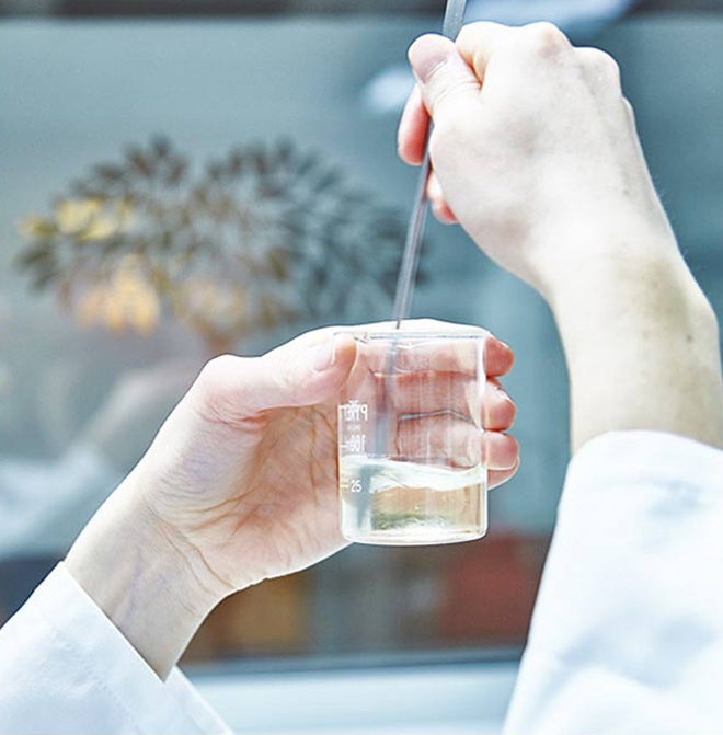 Close-up das mãos de uma pessoa segurando um copo de vidro contendo um líquido transparente, com uma pipeta sendo usada para retirar o líquido. Uma silhueta desfocada de uma planta é visível ao fundo.