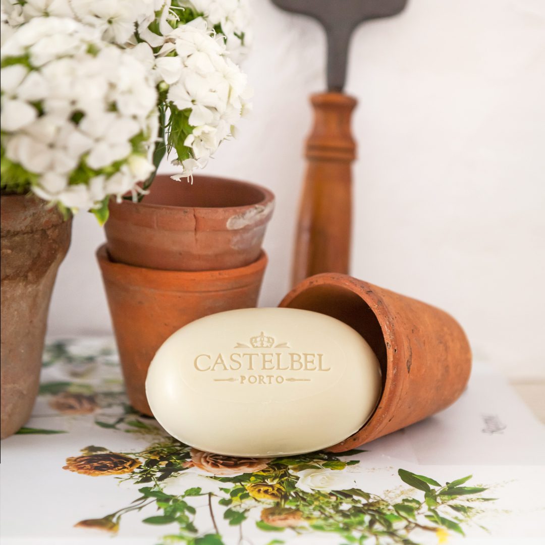 Un vaso di terracotta rovesciato con al suo interno un sapone CASTELBEL Porto, circondato da vasi di terracotta più piccoli e da una composizione floreale.