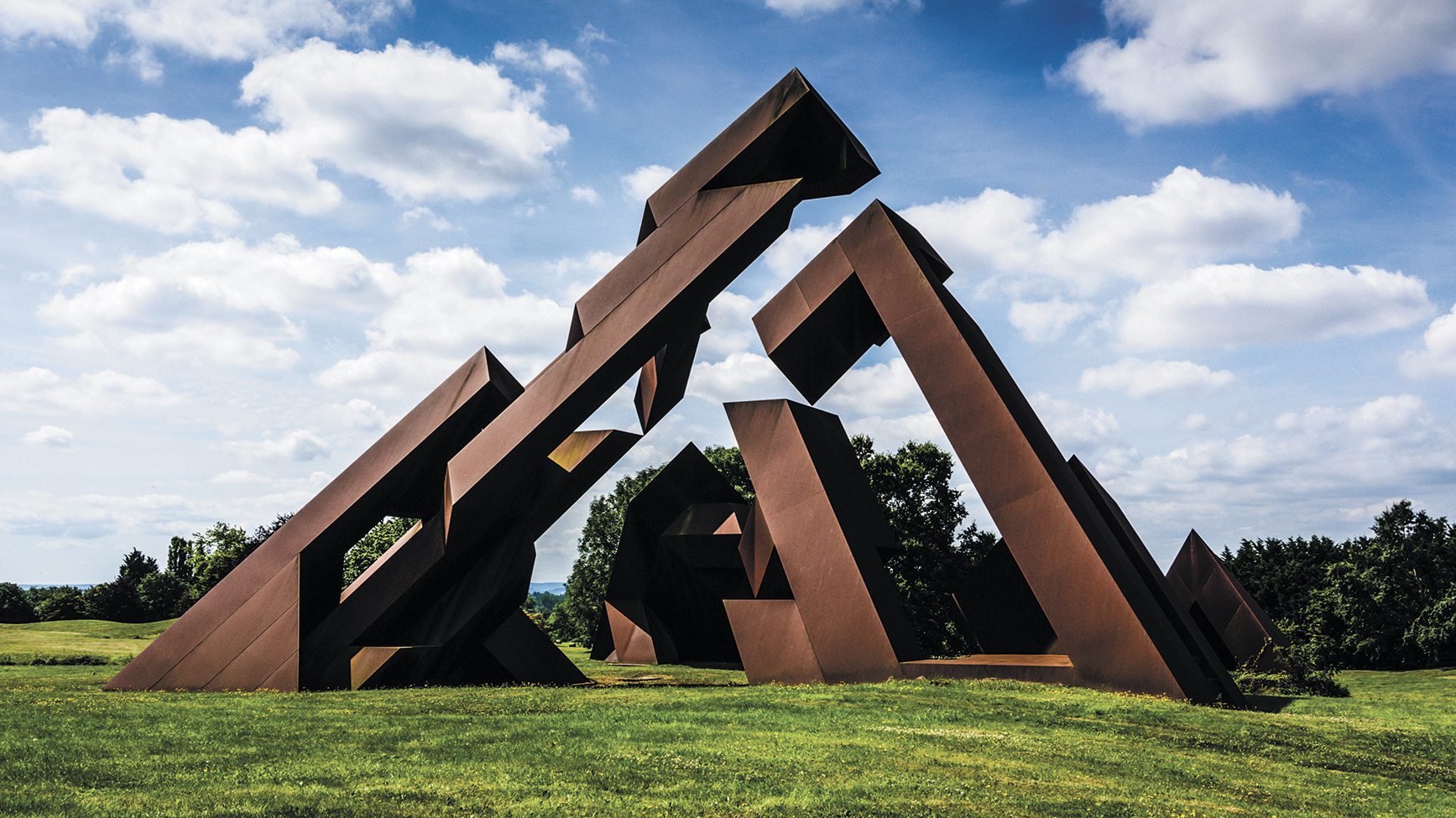Eine große, abstrakte Metallskulptur, die an kantige Gipfel erinnert, steht auf einer grünen Wiese unter einem bewölkten Himmel. Die Struktur zeichnet sich durch rostfarbene Oberflächen und dynamische geometrische Formen aus.
