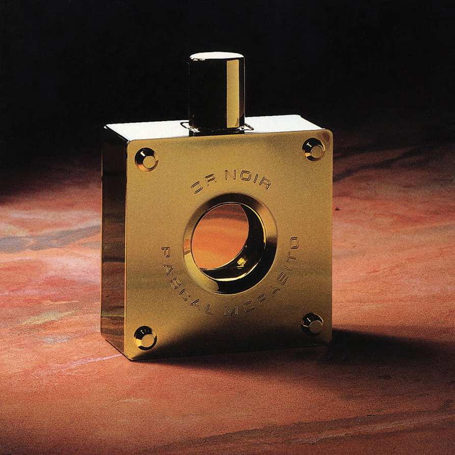 Ein goldener, rechteckiger Parfümflakon mit schlichtem Design und einer runden Öffnung in der Mitte, der auf einer strukturierten Oberfläche steht. Auf dem Flakon sind die Worte „De Noir“ und „Pascale Michelon“ eingraviert.
