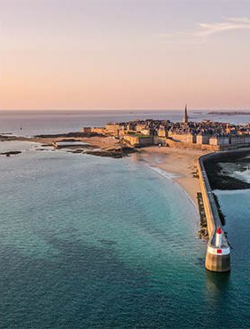 Een kustzicht van een stad bij zonsondergang, met een zandstrand, rotsachtige kustlijn en een vuurtoren aan het einde van een pier. De rustige zee weerspiegelt de warme kleuren van de lucht.