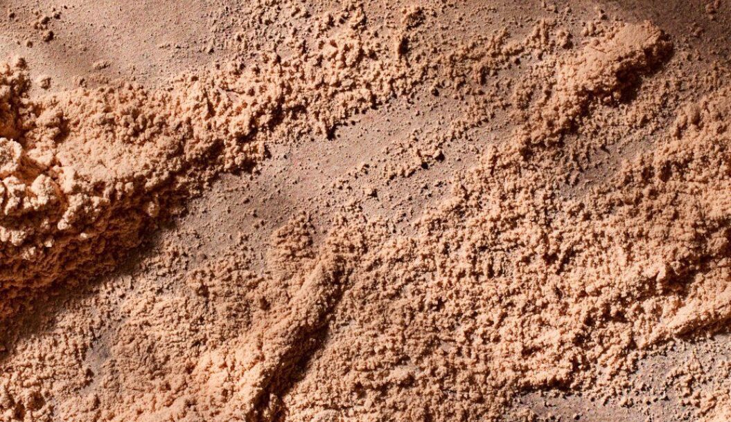 Uma imagem em grande plano de um pó castanho-avermelhado de textura fina, semelhante a areia ou terra, com superfícies irregulares e sombras subtis.