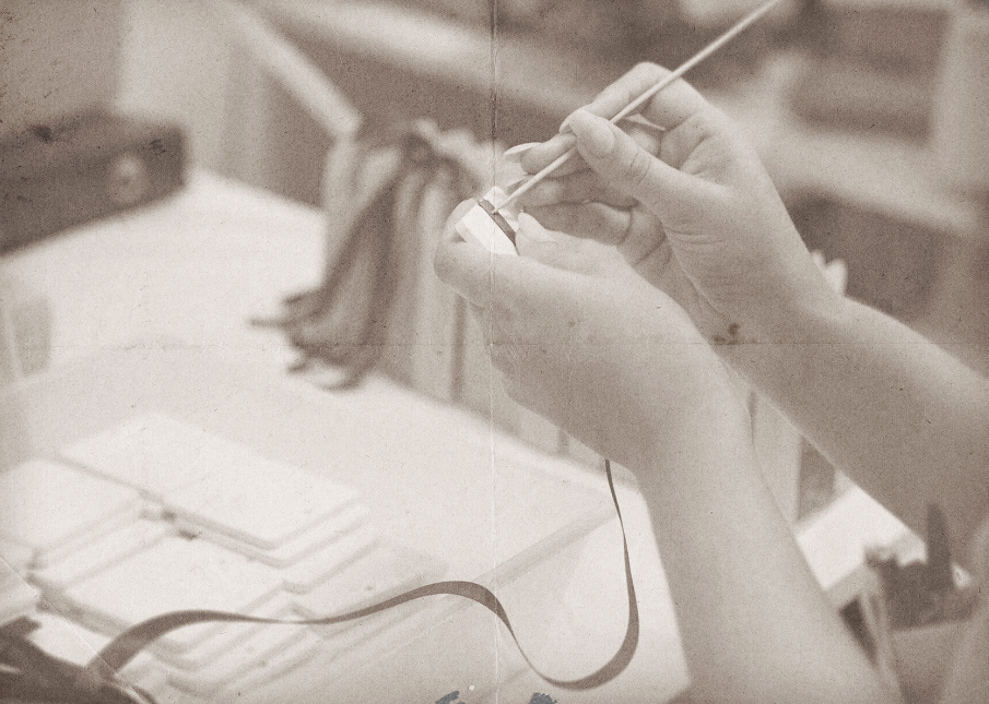 Le mani di una persona stanno dipingendo con cura un piccolo oggetto bianco mentre sono circondate da vari materiali artistici. L'immagine ha una tonalità vintage, color seppia.