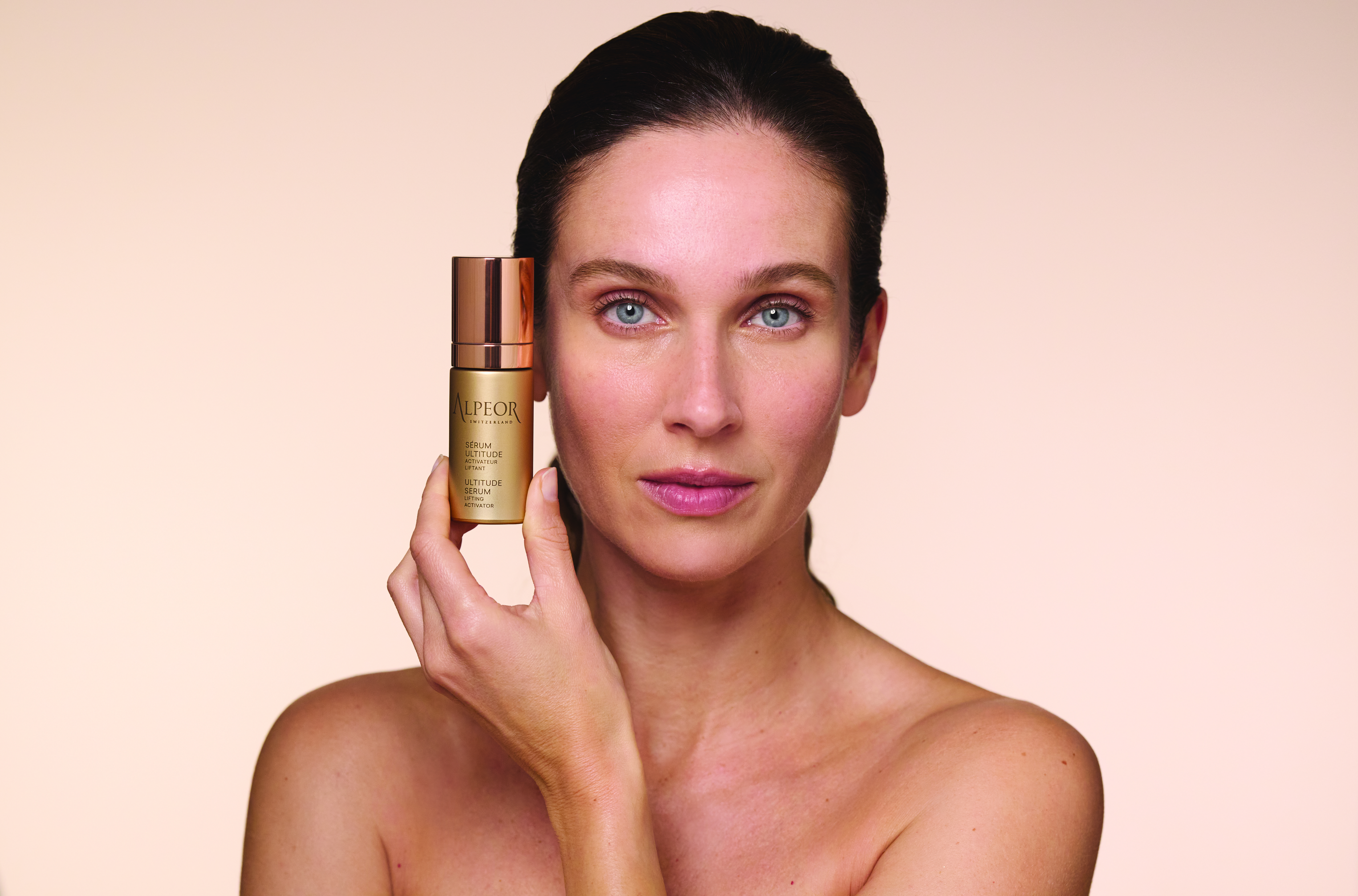 Uma mulher com cabelos longos e escuros segura um frasco de produto para a pele perto do rosto, mostrando a sua pele radiante contra um fundo suave e neutro.