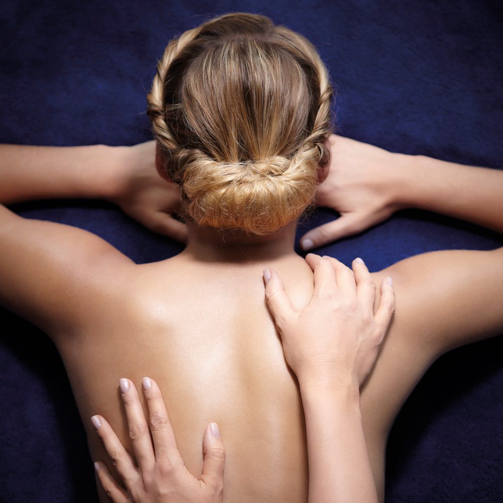 Een persoon met lang, gevlochten haar ligt op zijn buik en krijgt een rugmassage. Een andere hand oefent zachtjes druk uit op de schouderzone.