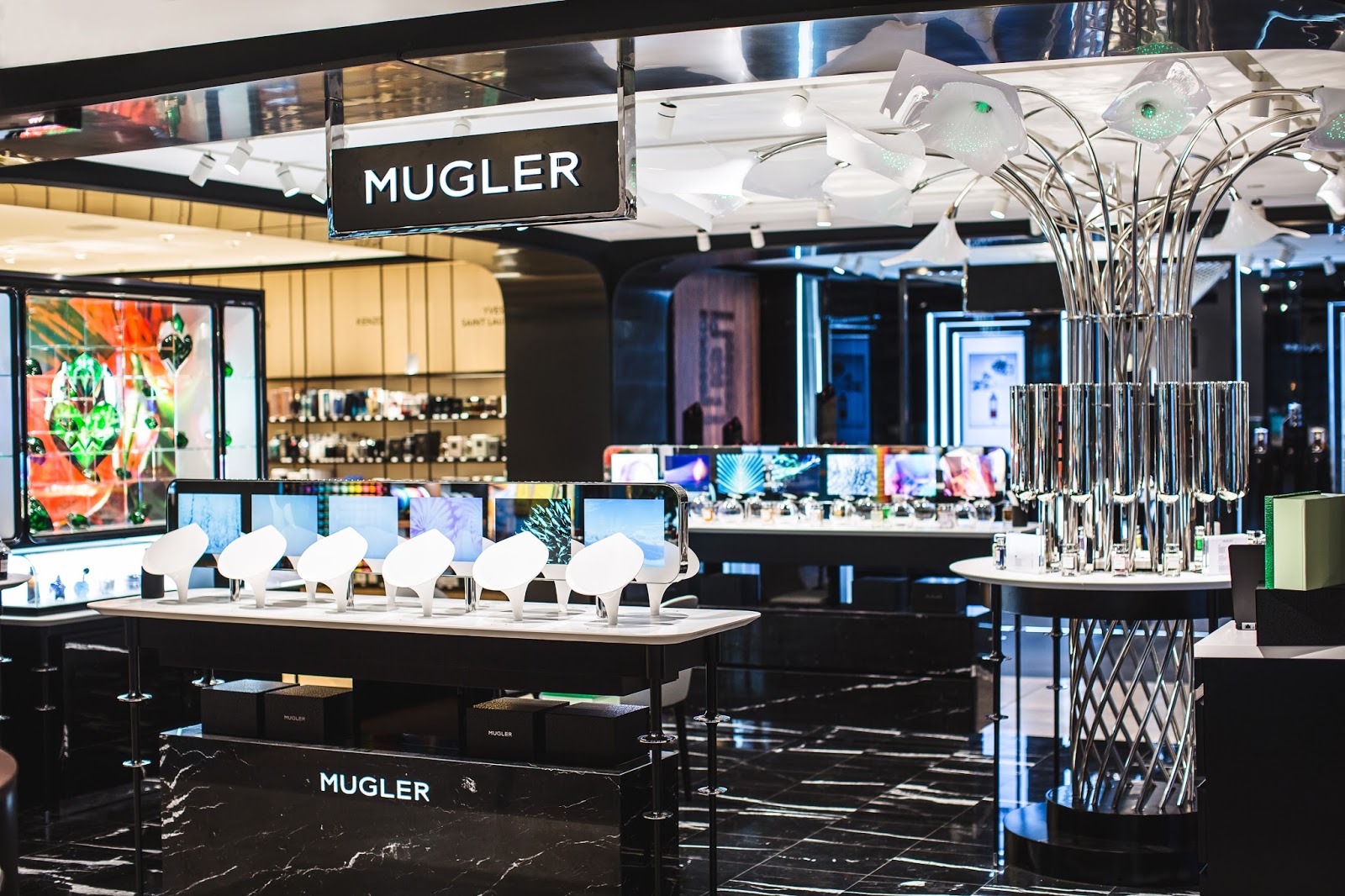 Şık siyah-beyaz vitrinleriyle dikkat çeken modern bir parfümeri; arka plandaki canlı sanat eserleri eşliğinde Mugler ait çeşitli kokuları sergiliyor.