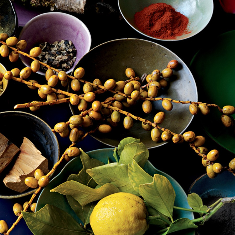 Uma coleção colorida de taças decorativas repletas de especiarias, ervas aromáticas e frutas coloridas, incluindo um limão e cachos de pequenas frutas amarelas, dispostas artisticamente sobre um fundo escuro.