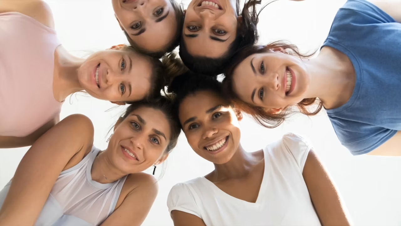 Um grupo de seis mulheres sorridentes está reunido, inclinado em direção à câmara, com um fundo claro e brilhante. Elas exibem expressões alegres e penteados variados.