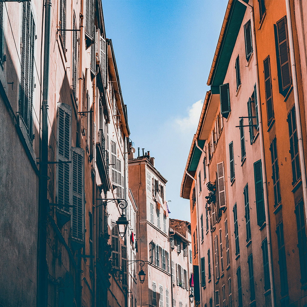 Una stradina stretta fiancheggiata da edifici alti, con un mix di facciate colorate e finestre con le persiane chiuse, sotto un cielo limpido e azzurro.