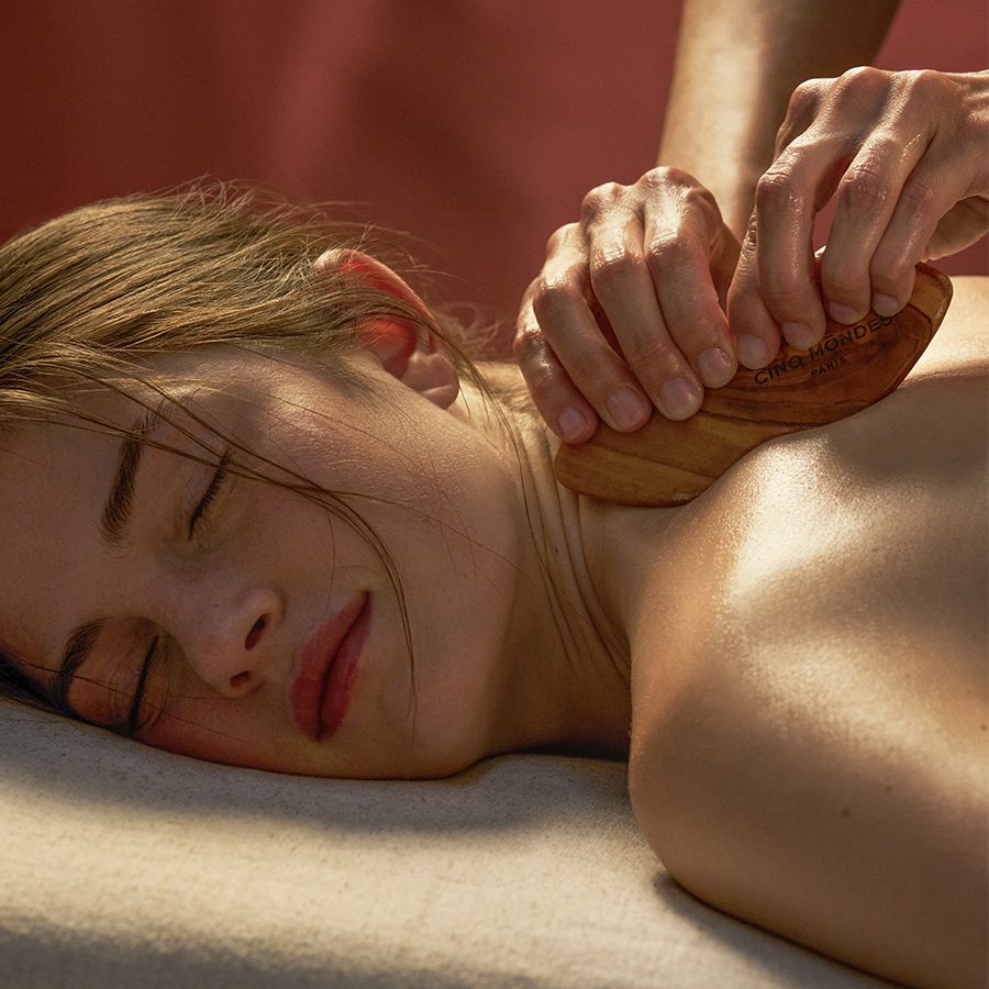 Uma pessoa recebe uma massagem nas costas com utensílios de madeira, enquanto está deitada numa superfície macia, de olhos fechados, com uma expressão relaxada. O ambiente é iluminado por uma luz quente e suave.