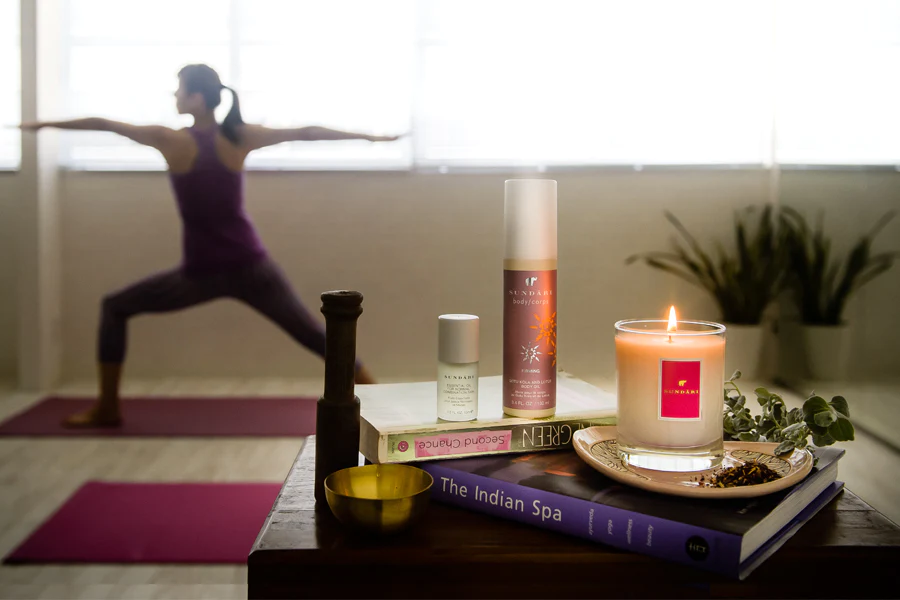 Um ambiente sereno de ioga com uma pessoa na postura do Guerreiro II ao fundo, com produtos para a pele, uma vela acesa e livros expostos em primeiro plano. A atmosfera sugere relaxamento e bem-estar.
