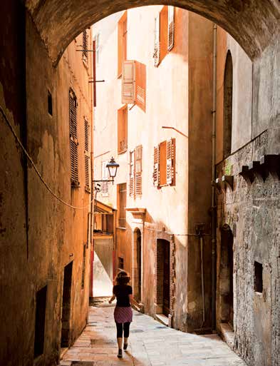 Una persona cammina lungo uno stretto vicolo illuminato dal sole, con vecchi edifici e persiane di legno che costeggiano le pareti. L'arco sopra la sua testa incornicia la scena, creando un'atmosfera pittoresca.