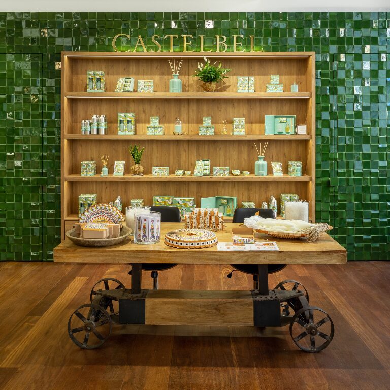 Uma mesa de exposição em madeira em frente a uma parede revestida com azulejos verdes exibe vários produtos Castelbel, incluindo sabonetes, velas e artigos decorativos, dispostos numa prateleira de madeira atrás.