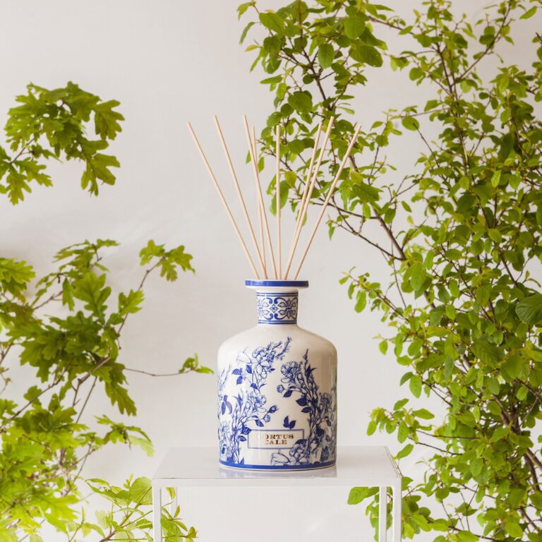 Um difusor decorativo de cerâmica azul e branca repousa sobre um suporte transparente, com várias varas de bambu a emergir do seu gargalo, tendo como pano de fundo uma folhagem verdejante.