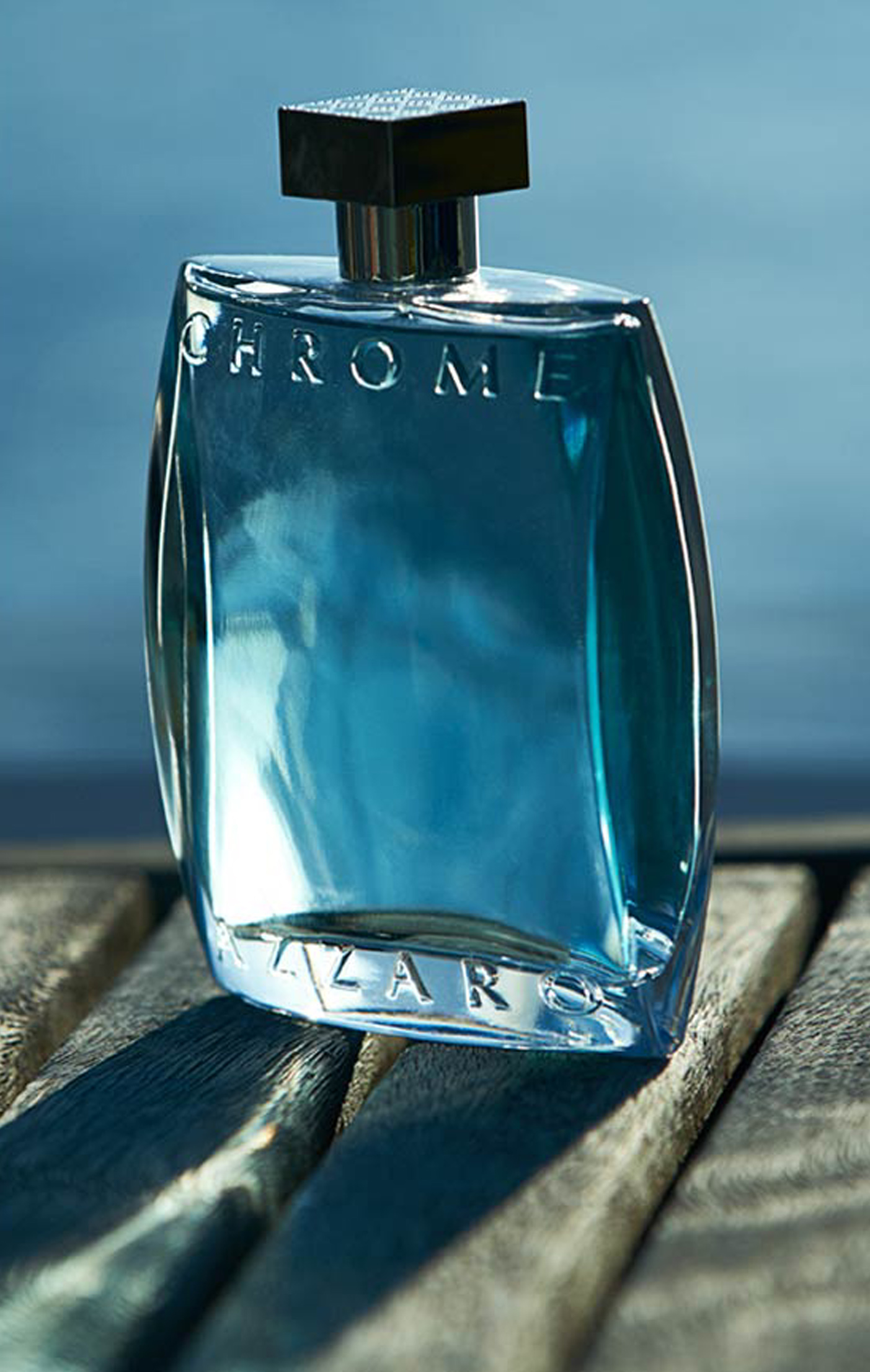 A clear glass bottle of Chrome by Azzaro perfume, with a blue liquid inside, placed on a wooden surface. The background features a blurred blue color, suggesting water or sky.