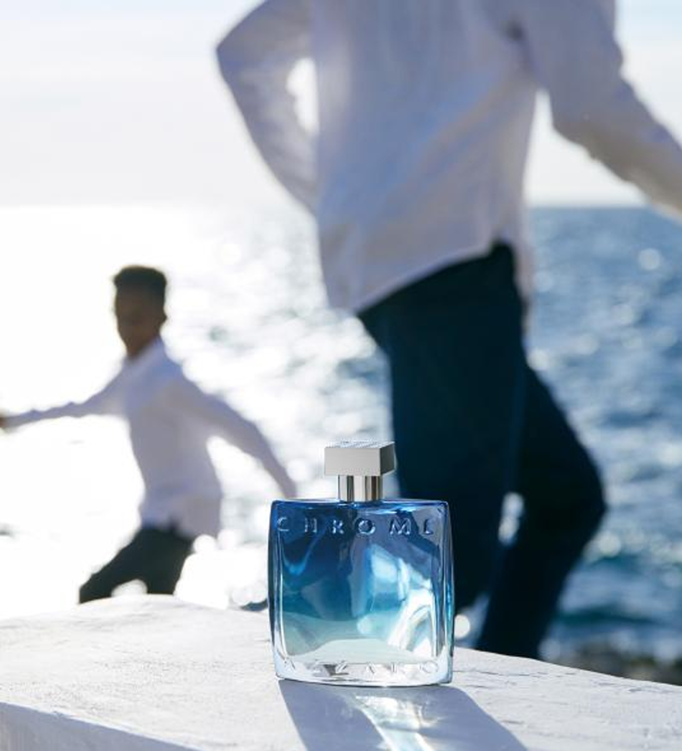 A bottle of blue perfume sits on a ledge with a blurred figure in a white shirt playfully running along the shore in the background, set against a shimmering ocean.